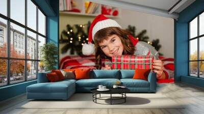 cute little happy smiling schoolgirl 8 years old wearing a santa hat and sitting at home on the bed holding a gift christmas and new year theme Wall mural