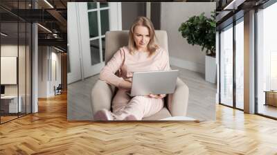 A beautiful blonde middle-aged woman sees in a cozy home chair with a laptop in her hands. Work or study from home. Freelancing or distance learning. Wall mural