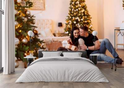 Young couple man and girl at home sitting by the Christmas tree, in the new year or christmas. Give gifts to each other. Wall mural