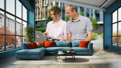 Attractive young woman and senior man are using laptop sitting on the bench in modern residential complex. Casual meeting outside office. A daughter is teaching technology for her father Wall mural