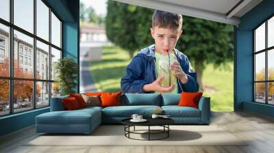 Boy 7-8 years old drinks lemonade from a plastic glass in the park and looks at the camera Wall mural