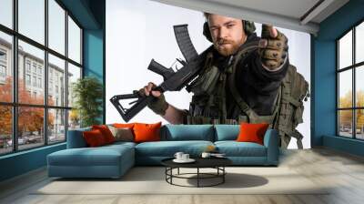 the airsoft player puts his rifle on his shoulder and aims at us with his fingers. a man in an outfit, in headphones, a bulletproof vest, with a backpack on a white background. Wall mural