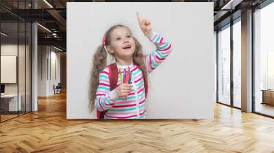 Back to school. Portrait of a blonde schoolgirl in a jacket with colored gel pens in her hands. The girl smiles and looks up. Thumbs up. Light background. Education. copy place. Wall mural