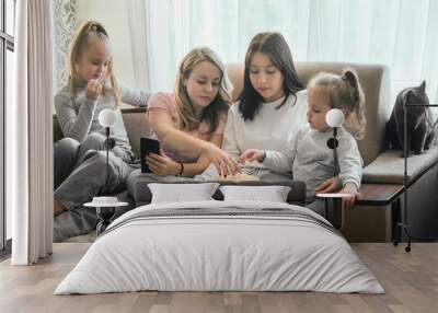 Two girls and two children have fun in a warm home environment, with a phone, watching TV and eating popcorn. a cat is sitting with them on the couch Wall mural