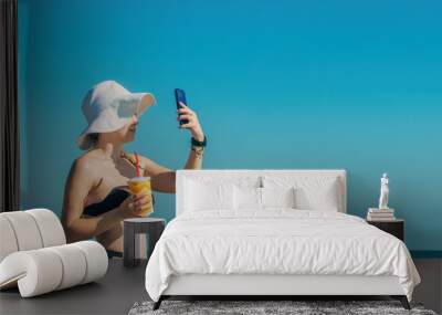 Young beautiful female tourist on vacation by the ocean in a sun hat and swimsuit, holds a glass with a cool summer drink in her hands, takes a selfie on the phone .copy space Wall mural