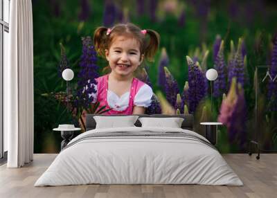 A small cheerful girl with two light tails on her head in a green field with purple flowers. A girl in a pink dress in a lupine field and a bouquet of lupines in her hands. Child's emotions-joy, prank Wall mural