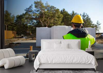 The builder  talking on the phone. Architect in a protective yellow helmet and a signal vest stands backs in front of the construction site of a house with a foundation and aerated concrete blocks.  Wall mural