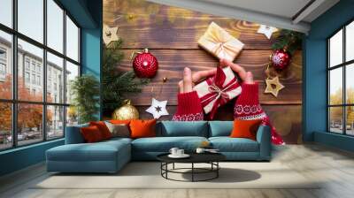 Women's hands in knitted sweater with ornament hold a box with Christmas gift in their palms on the table among the decor. New year, packaging and preparation of gifts, branches of fir, decoration Wall mural