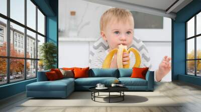 Cute caucasian blonde baby girl about 1 year old,sitting at chair having lunch,meal at kitchen,infant,holding whole banana.Fresh healthy infant food concept.Kid eating fruit.Neutral colors Wall mural