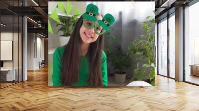 A girl with dark hair in funny green glasses, dressed in a green t-shirt, smiles looking at the camera, celebrating St. Patrick's Day at home. Wall mural