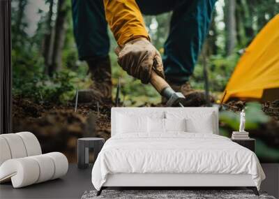 A low-angle view of someone hammering tent stakes into the ground, with the background softly blurred for action. Wall mural