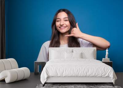 Attractive caucasian or arab brunette girl in a white t-shirt showing a call gesture on a mobile phone isolated in on a blue studio background. Wall mural