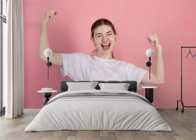 Attractive Caucasian young brunette woman in a casual white t-shirt screams with happiness and shows a winning gesture isolated on a pink studio background. Wall mural