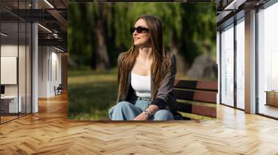 Attractive caucasian young woman in sunglasses and stylish casual clothes sitting on a park bench on a warm spring day. Rest, freedom, leisure. Wall mural