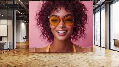 A cheerful young woman with curly hair wearing yellow sunglasses against a vibrant pink background during a sunny day Wall mural