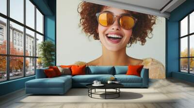 Bright and cheerful young woman with curly hair and stylish sunglasses enjoying a sunny day indoors while smiling broadly Wall mural