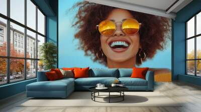 Joyful woman with curly hair smiles brightly while wearing stylish sunglasses against a sunny beach backdrop Wall mural