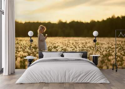 A little girl with blond hair in a white dress is picking flowers in a huge endless field of white fluffy dandelions. The sun is setting behind the forest. Image with selective focus. Wall mural