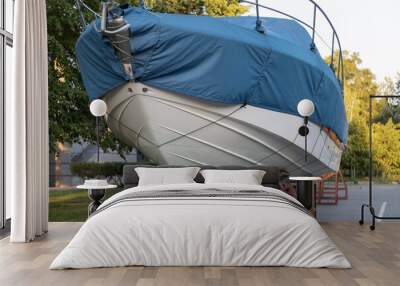 A motor boat covered with a blue cover stands on land under the open sky against the background of trees Wall mural