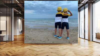 Two twins stand by the sea. Children in the same clothes are looking at the sea. Brothers boys in hats on the beach . Concept: vacation, friendship, brothers Wall mural