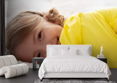 a beautiful girl with long blonde hair and beautiful eyes in a yellow jacket lies on a white bed and smiles Wall mural