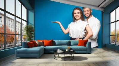 Cheerful business couple standing isolated over blue background, pointing fingers at copy space. Wall mural