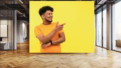 Cheerful smiling man points to a place for text on a yellow background, he is wearing an orange T-shirt, good news. Indian man Wall mural