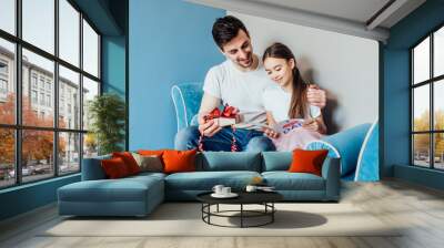 It is for you, dad!  Portrait of young man at home with his little cute girl, she is made present for dad. Happy Father's Day! Sitting on sofa at home. Wall mural