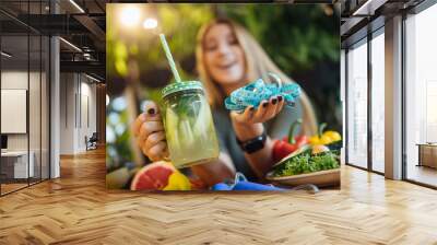 Photo of blonde fithess girl, trainer sit on the table and holding fresh smoothie and measure, diet time concept Wall mural