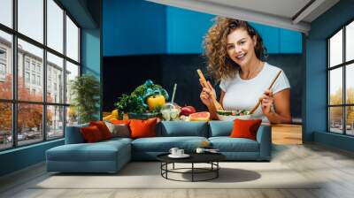 Young and happy woman eating healthy salad sitting on the table with green fresh ingredients indoor. Wall mural