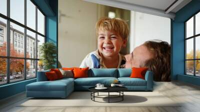 A smiling young boy sitting on his father's lap, both sharing a joyful and playful moment together at home.
 Wall mural