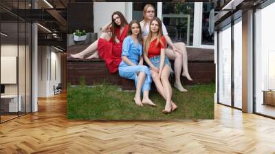 Group of young confident good-looking barefoot women with long hair sitting on wooden veranda near green grass, summer. Wall mural