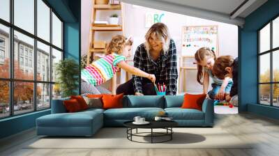 portrait of young joyful woman teacher sit with children, looking at serious little girl taking penc Wall mural