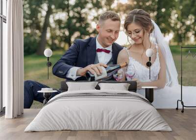 Smiling and shy groom and bride drinking and pouring bubbly champagne wine from bottle to glasses together, sit in park Wall mural