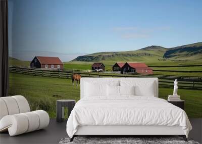 Two icelandic horses grazing on a green pasture in front of a traditional icelandic farm with red roofs and wooden fences Wall mural