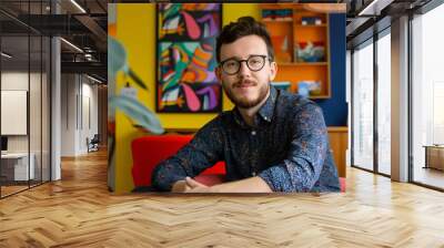 Young creative professional with a beard and glasses is sitting in a colorful office space, looking relaxed and confident Wall mural