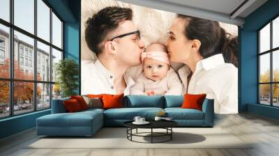 Happy young father, mother and cute baby boy lying on light bed. Mother and father kiss baby. Wall mural