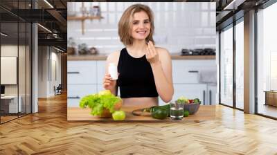 Beauty supplement. Attractive young woman holding vitamin capsule and glass of  water  at the kitchen. Healthy lifestyle, healthy diet nutrition concept Wall mural