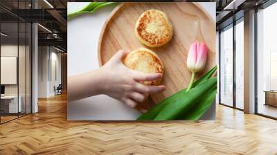 Closeup fresh pink tulip near wooden round plate, the hand of child takes two golden bitten fried cheesecakes in the shape of figure 8, a dessert made from tasty cottage cheese Wall mural