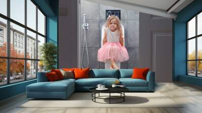 Cute little girl in white pink dress sitting on toilet with toilet paper on background of walls with gray tiles, view from open door. Wall mural