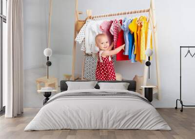 Baby girl stands near a wardrobe and chooses a dress and smiles. Dressing closet with clothes arranged on hangers. Colorful wardrobe of newborn, kids, toddlers, babies full of all clothes Wall mural