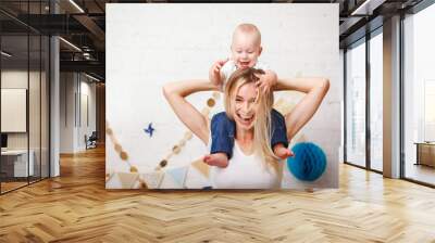 A cute handsome one-year-old baby sitting by her mother's neck grabs her by the hair and laughs. A beautiful young mother with her son play laughing and look at the camera. Mothers Day. Close-up Wall mural