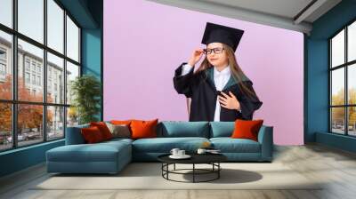 A beautiful graduate in a black graduation ceremonial robe looks thoughtfully into the distance and holds her glasses. isolated purple background. Wall mural