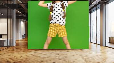 A full-length teenage girl dressed in a summer hat and shorts. A beautiful little lady holds her head in her hands and looks up. Summer clothes for recreation. Green isolated background. Wall mural