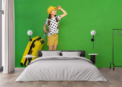 A little girl with a suitcase on wheels looks into the distance. A child in shorts and a hat is eating a vacation at the sea. Green isolated background. Copy space. Wall mural