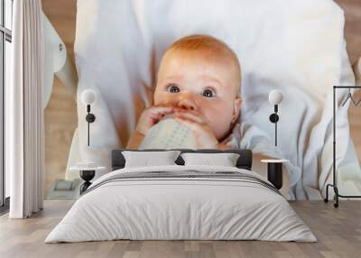 Cute little newborn girl drinking milk from bottle and looking at camera on white background. Infant baby sucking eating milk nutrition lying down on crib bed at home. Motherhood happy child concept Wall mural