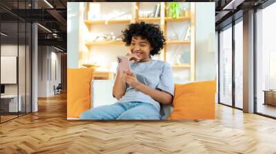 Excited happy young black african american woman holding cell phone laughing feeling joy getting mobile message. Overjoyed girl laughing aloud sitting on couch watching funny video reading news Wall mural