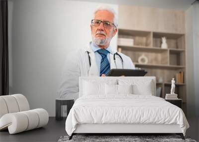 Mature senior male doctor in medical uniform using digital tablet in clinic. Therapist professional healthcare expert searching information online in hospital room. Medicine healthcare medical checkup Wall mural