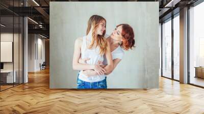 Stay at home mom, stay safe. Young mother embracing her child. Woman and teenage girl relaxing in white bedroom near gray wall indoors. Happy family at home. Young mom playing with her daughter. Wall mural