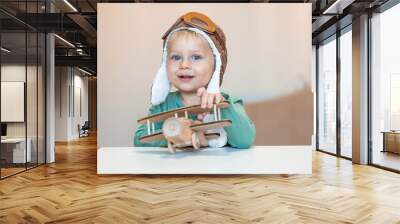 A handsome little boy of 2 years old in a pilot's hat is playing with a wooden airplane and a logistics constructor. Children's wooden toys for child development. Wall mural